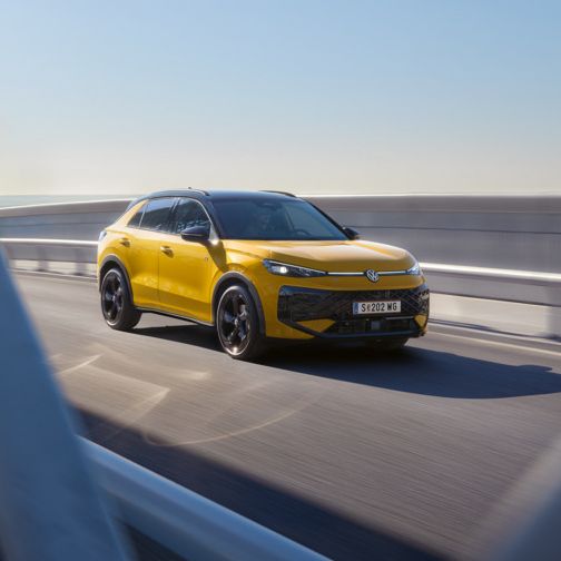 Ein gelber T-Roc fährt über eine moderne Brücke bei klarem Himmel. Die Aufnahme zeigt das Fahrzeug aus der Ferne, teilweise durch die Brückenstrukturen hindurch. Es ist ein sonniger Tag.