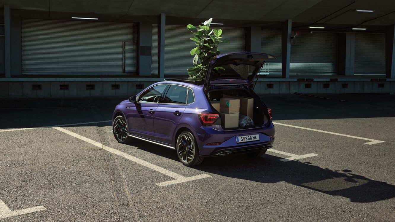 Lila VW Polo mit offener Heckklappe und Pflanze im Schiebedach vor Lagerhalle.