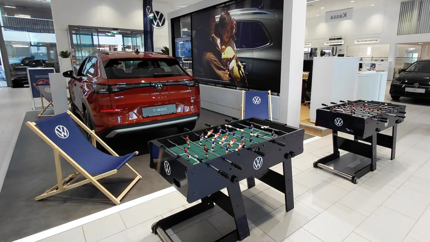 Innenraum eines VW-Autohauses mit einem roten Elektroauto (VW ID.4) auf einer Präsentationsfläche neben einem blauen Liegestuhl mit VW-Logo. Im Vordergrund stehen zwei Kickertische, ebenfalls mit VW-Logo, die einen Freizeitbereich für Kunden andeuten. Im Hintergrund sind weitere Fahrzeuge, Verkaufsflächen und Bildschirme zu sehen.