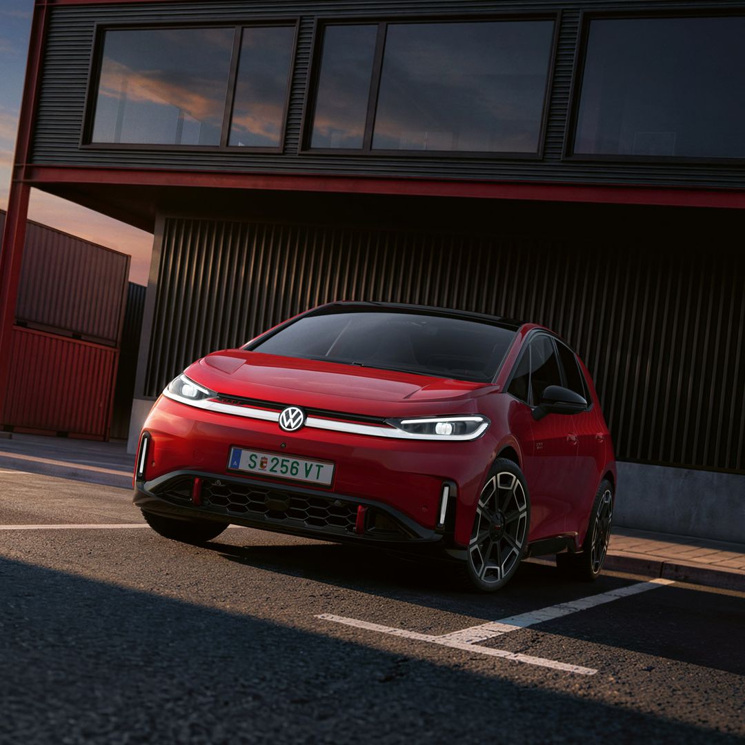 [AI-modified] – Frontansicht eines roten VW ID.3 GTI mit eingeschalteten Scheinwerfern und beleuchtetem Logo vor einem Gebäude.