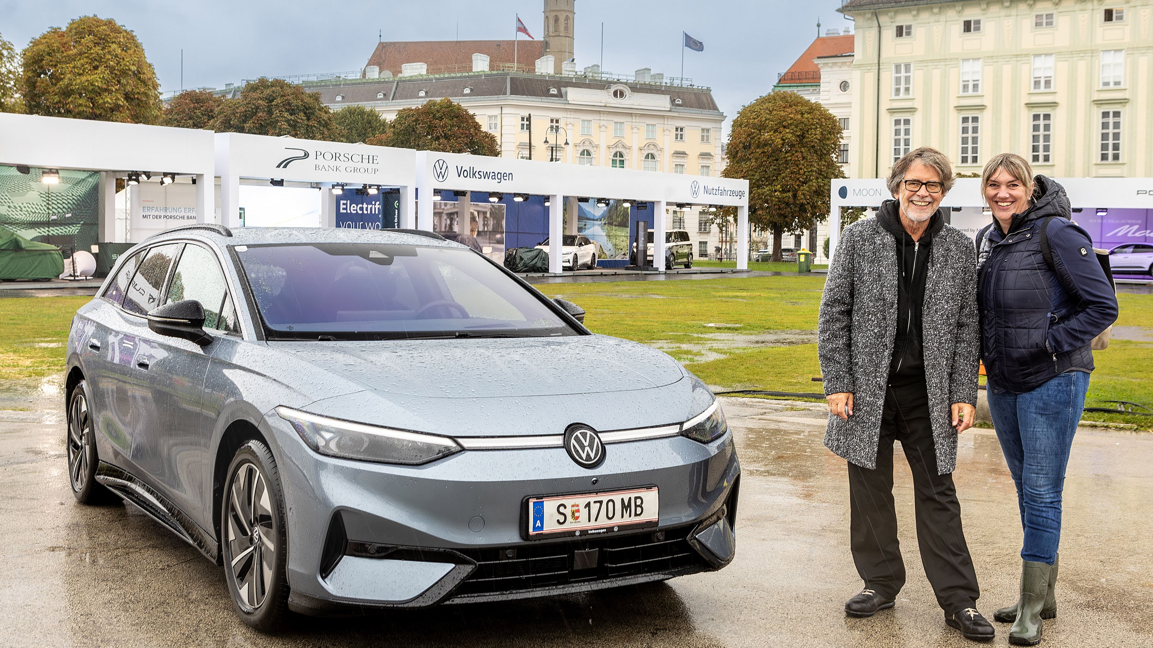 Zwei Personen stehen neben einem silbergrauen Volkswagen ID.5 Elektroauto auf einem Messegelände mit verschiedenen Ausstellungsständen im Hintergrund.