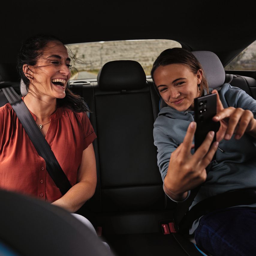 2 Peronen sitzen auf der Rückbank eines Volkswagen und schauen lächelnd auf ein Mobiltelefon