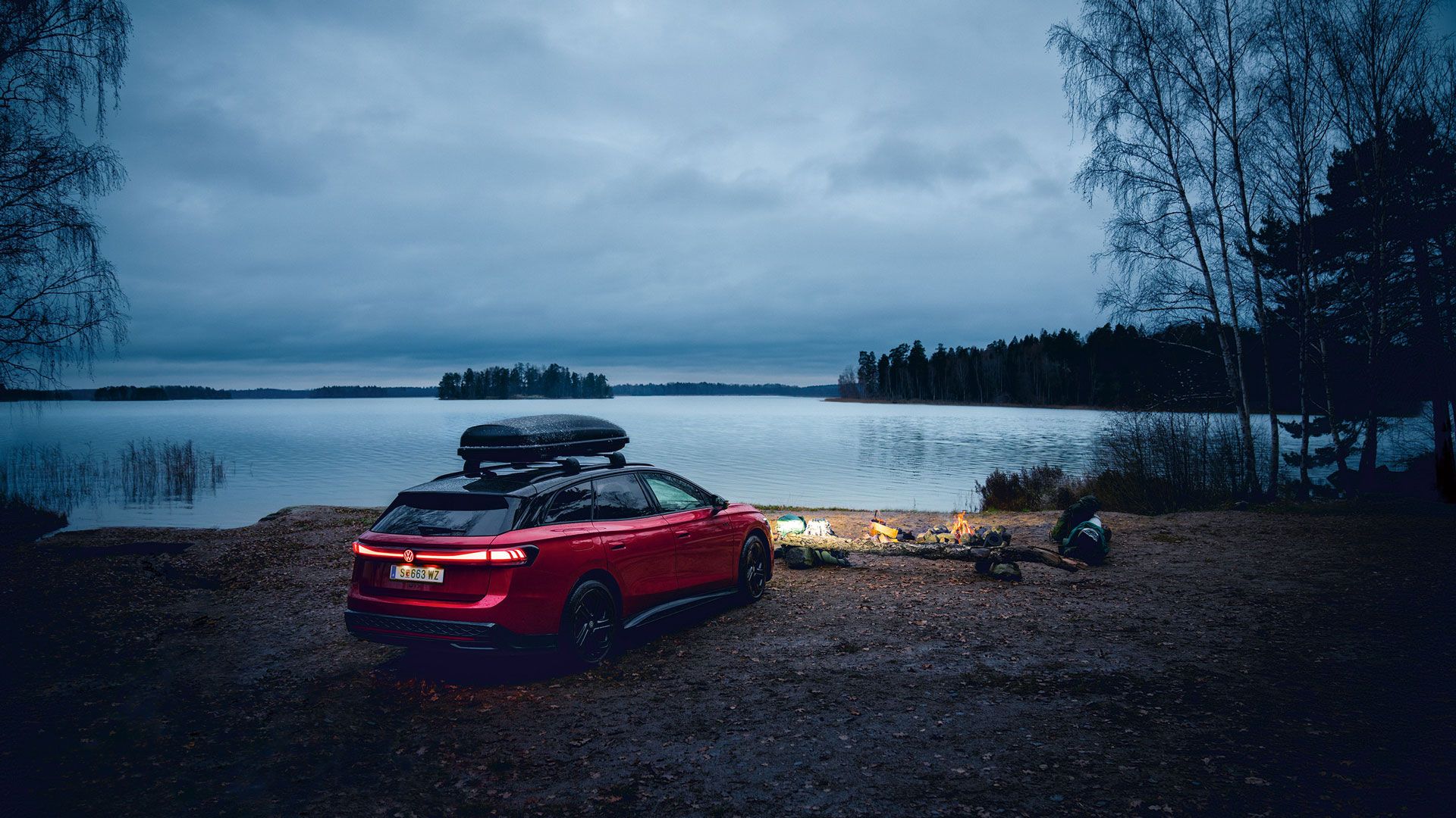 Roter VW ID.7 GTX Tourer mit Dachbox parkt an einem See in der Dämmerung.