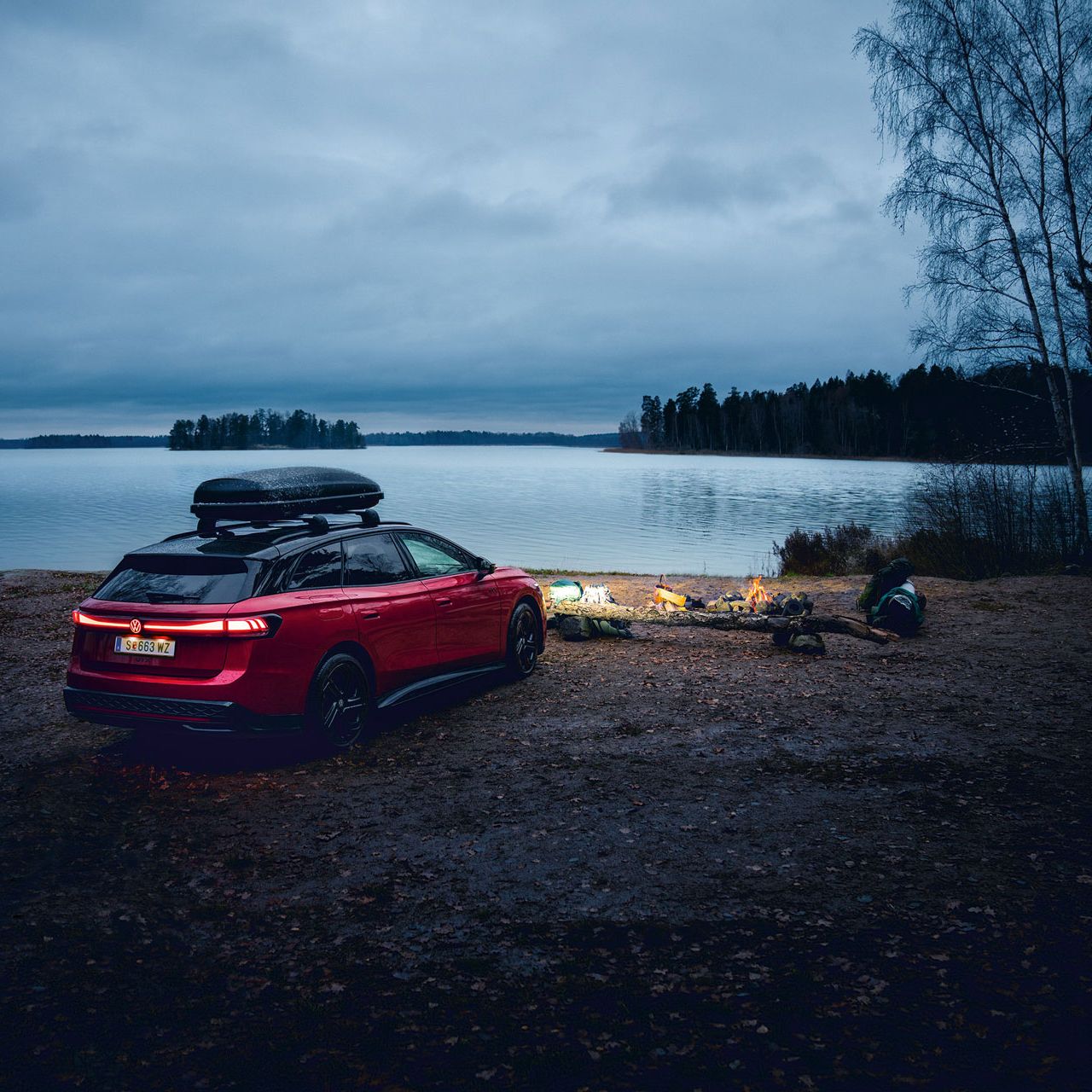 Roter VW ID.7 GTX Tourer mit Dachbox parkt an einem See in der Dämmerung.