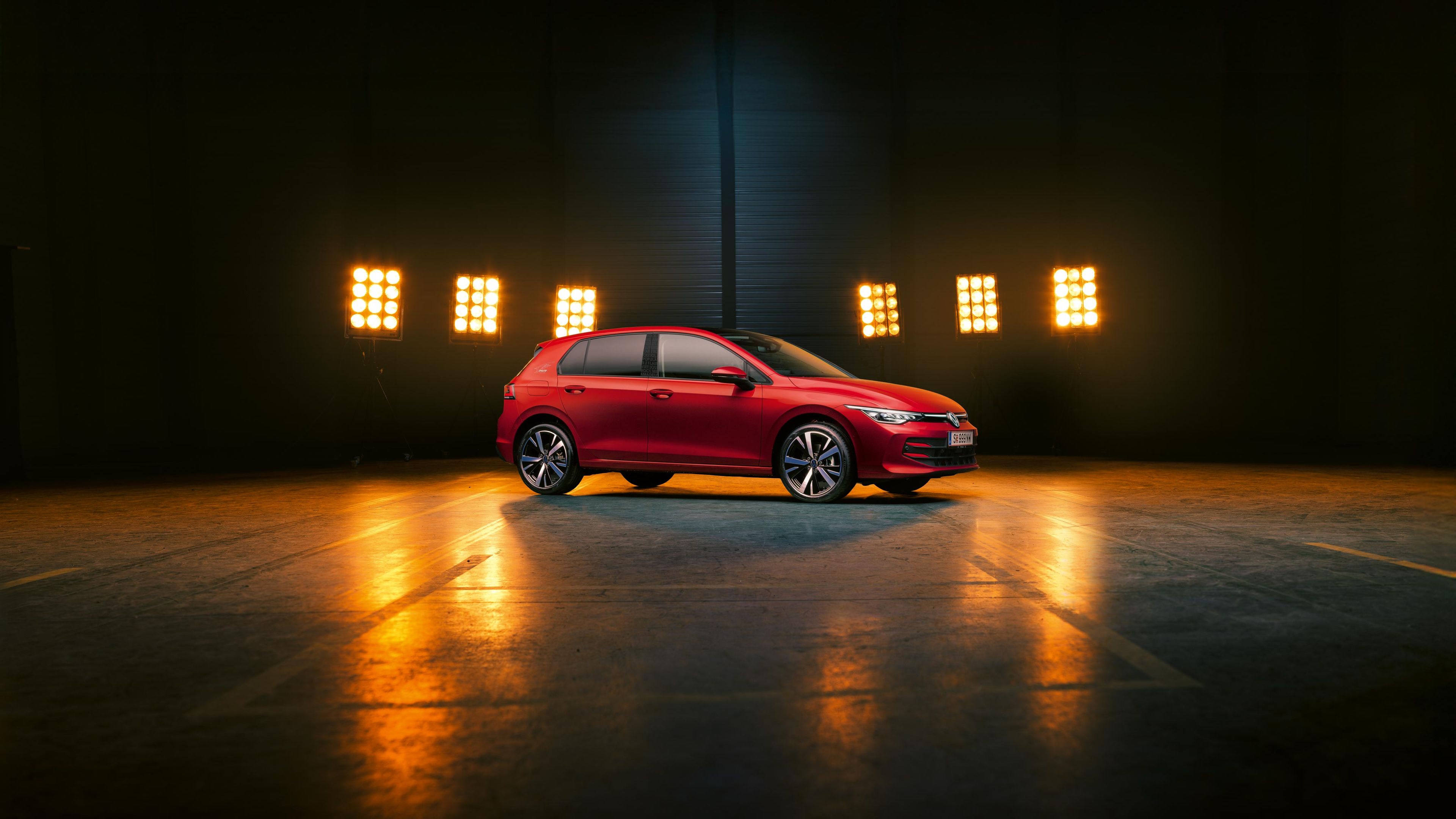 Seitenansicht auf einen roten VW Golf Falco Edition, der in einer leeren Halle steht, dahinter wird er von Scheinwerfern beleuchtet.