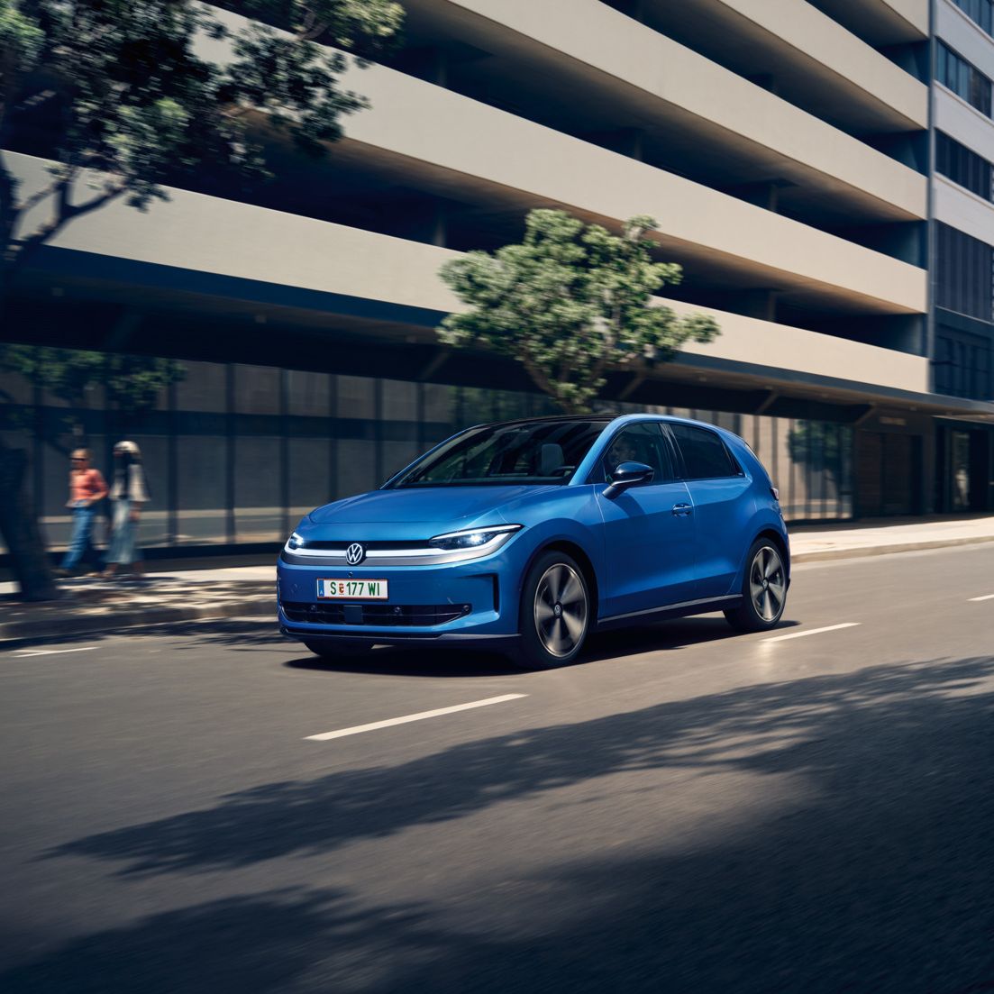 Ein blauer VW ID. Polo Style fährt durch eine Stadt mit grünen Bäumen am Straßenrand.