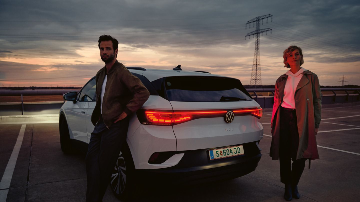 Frau und Mann stehen neben dem Heck eines weißen VW ID.4 bei Dämmerung auf einem Parkplatz
