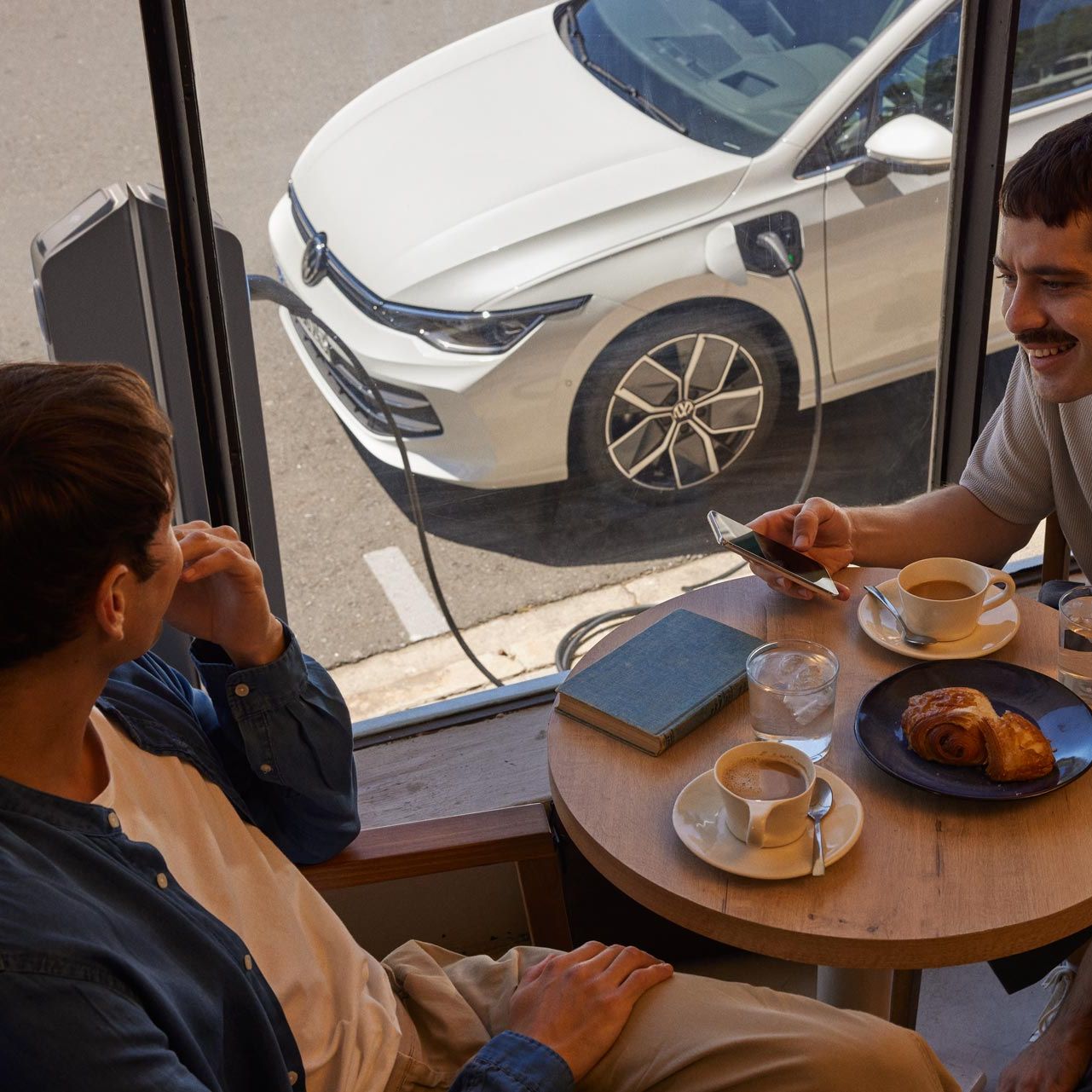 Zwei Männer sitzen in Café und unterhalten sich, durch das Fenster sieht man einen VW Golf eHybrid der geladen wird
