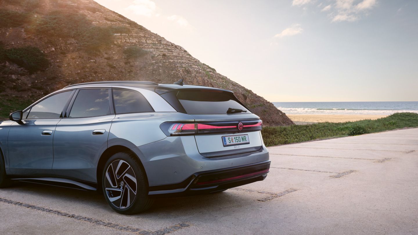 Die Seitenansicht auf das Heck eines blauen VW ID.7 Tourer, geparkt auf einem Parkplatz nahe eines Bergs, im Hintergrund ein Strand und das Meer.