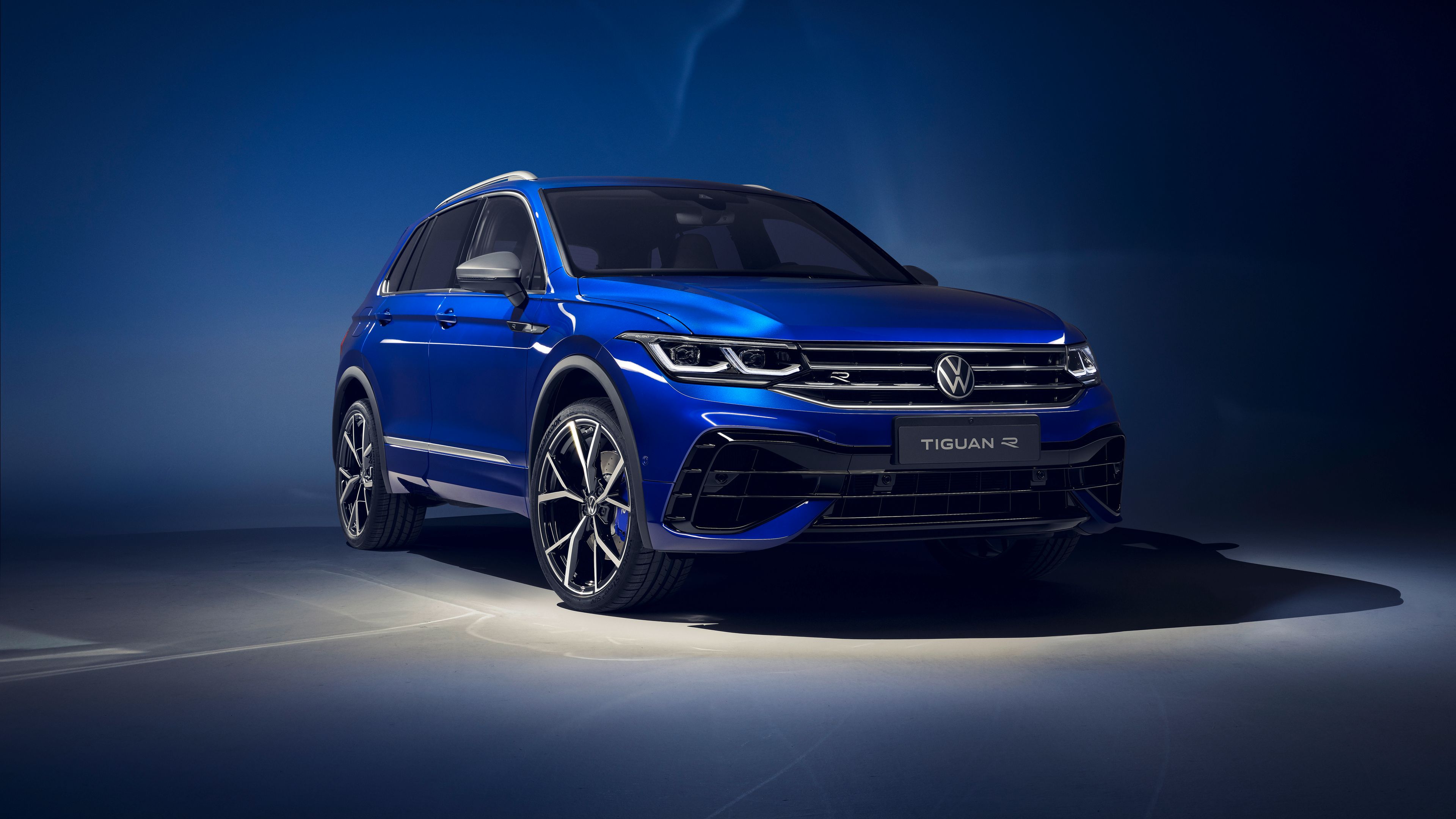Ein blauer Volkswagen Tiguan R steht vor einem dunklen Hintergrund. Das Auto hat sportliche Merkmale und große Felgen. Der Schriftzug "Tiguan R" ist auf einem Schild unter dem Kühlergrill sichtbar.