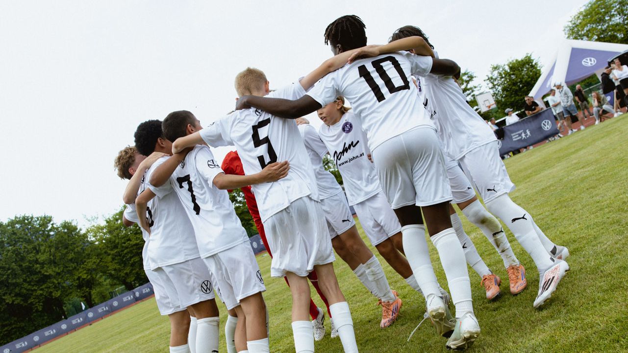 Eine Jugendfußballmannschaft in weißen Trikots steht im Kreis auf dem Spielfeld und legt die Arme umeinander. Die Spieler tragen Rückennummern, darunter gut sichtbar die Nummern 5 und 10. Im Hintergrund sind Bäume, ein Zaun mit Werbebannern und ein blauer Himmel mit ein paar Wolken zu sehen.