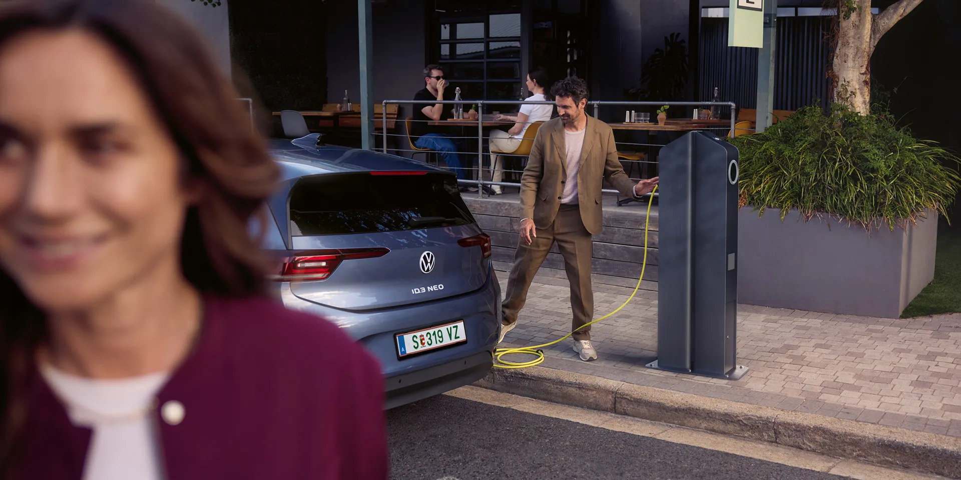 Eine Frau geht auf einer Straße. Hinter ihr läd ein Mann einen VW ID.3 Neo an einer Ladesäule vor einem Café