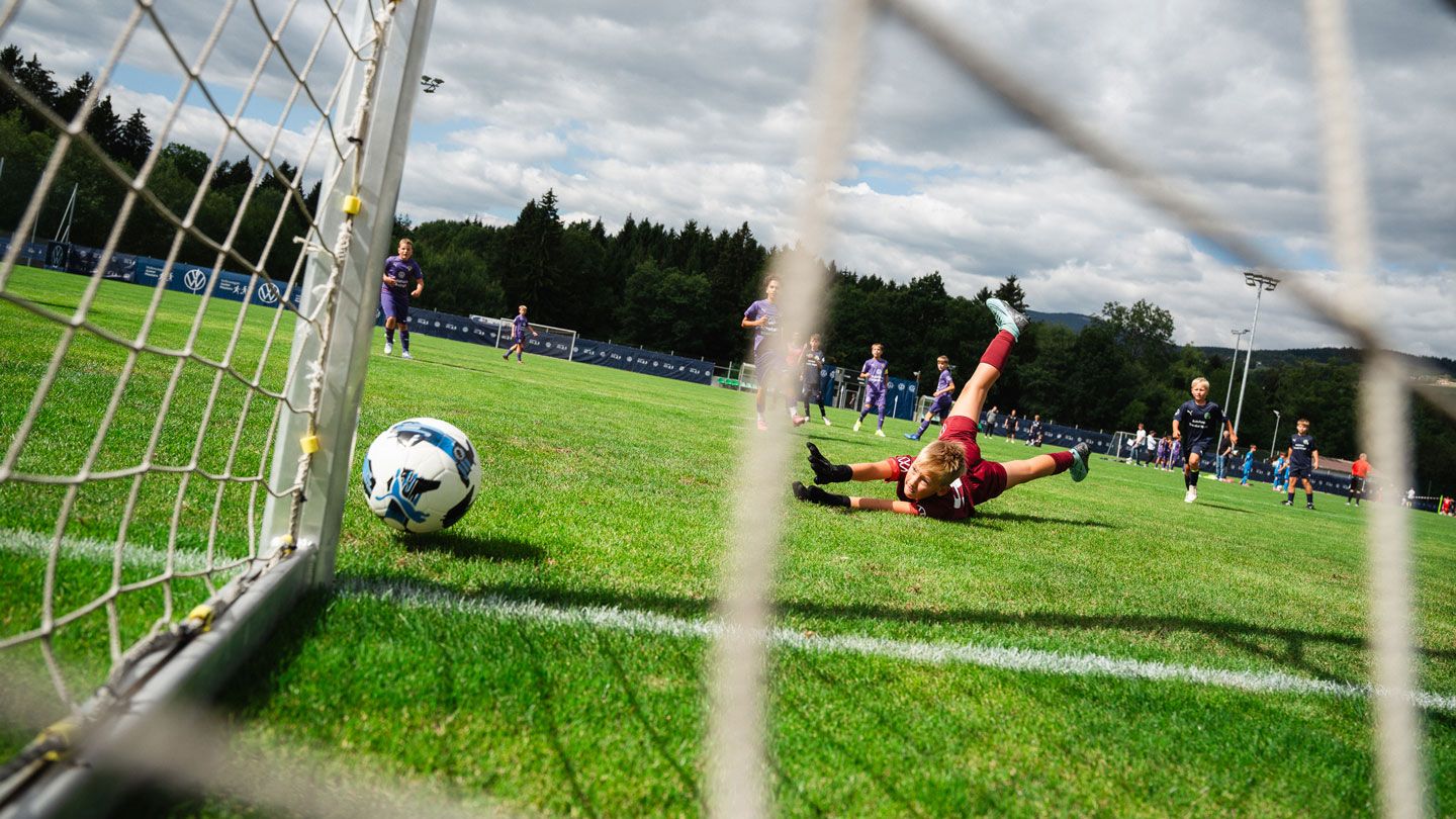 Ein Fußball rollt gerade Richtung Tor, während der Torwart vergeblich ausgestreckt auf dem Rasen liegt. Im Hintergrund sind weitere Kinder auf dem Spielfeld zu sehen, die an einem Spiel auf einem großen, grünen Rasenplatz teilnehmen. Der Himmel ist leicht bewölkt und im Hintergrund erkennt man Bäume und Flutlichtmasten.