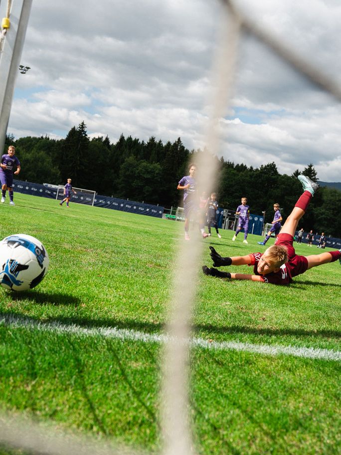 Ein Fußball rollt gerade Richtung Tor, während der Torwart vergeblich ausgestreckt auf dem Rasen liegt. Im Hintergrund sind weitere Kinder auf dem Spielfeld zu sehen, die an einem Spiel auf einem großen, grünen Rasenplatz teilnehmen. Der Himmel ist leicht bewölkt und im Hintergrund erkennt man Bäume und Flutlichtmasten.