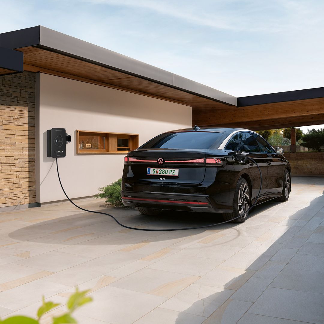 Ein schwarzes Elektroauto steht in einer modernen Einfahrt und ist an einer Wallbox an der Hauswand zum Laden angeschlossen. Das flache, kubische Haus mit Holz- und Steinelementen wirkt modern und hochwertig. Im Vordergrund sind Pflanzen zu sehen, im Hintergrund öffnet sich der Blick auf eine Landschaft.