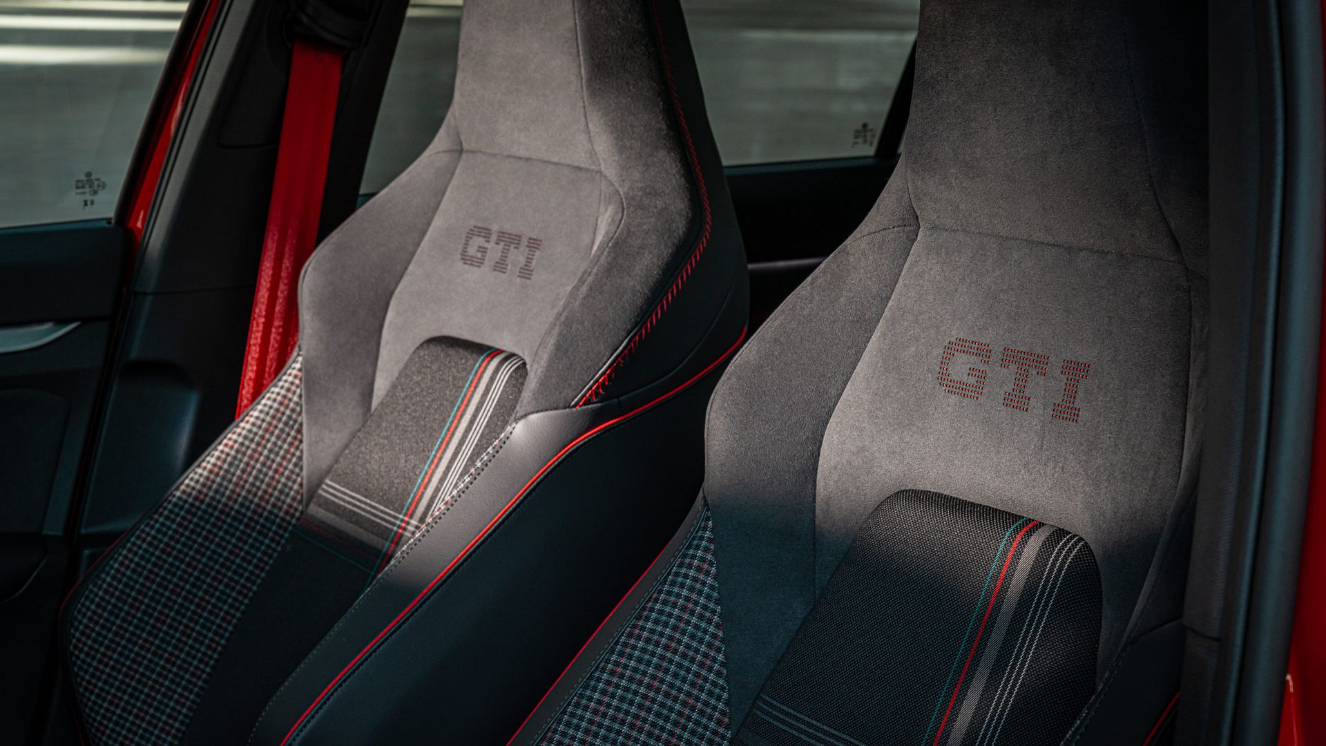 Zwei Autosportsitze mit gesticktem "GTI"-Logo. Die Sitze haben schwarze und rote Farbakzente sowie ein kariertes Muster in der Mitte. Sie befinden sich in einem Fahrzeug.