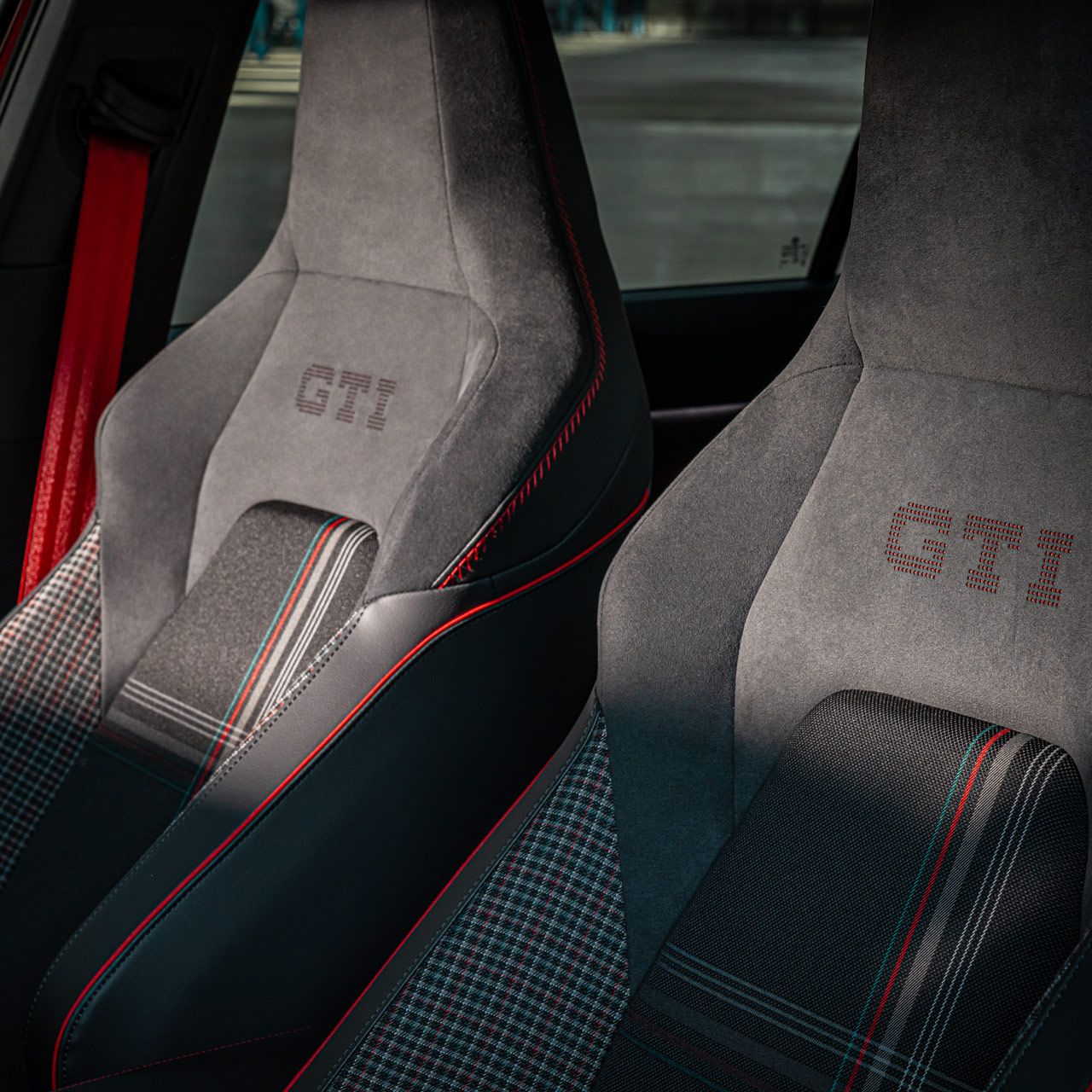 Zwei Autosportsitze mit gesticktem "GTI"-Logo. Die Sitze haben schwarze und rote Farbakzente sowie ein kariertes Muster in der Mitte. Sie befinden sich in einem Fahrzeug.
