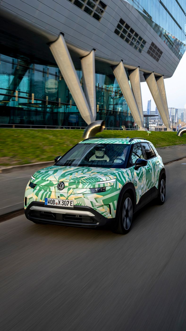 Ein Elektroauto fährt auf einer Straße neben einem modernen Gebäude mit schrägen Stützen und großer Glasfassade. Das Auto hat eine grüne und weiße Tarnlackierung. Im Hintergrund sind weitere Gebäude und eine graue, bewölkte Skyline zu sehen.