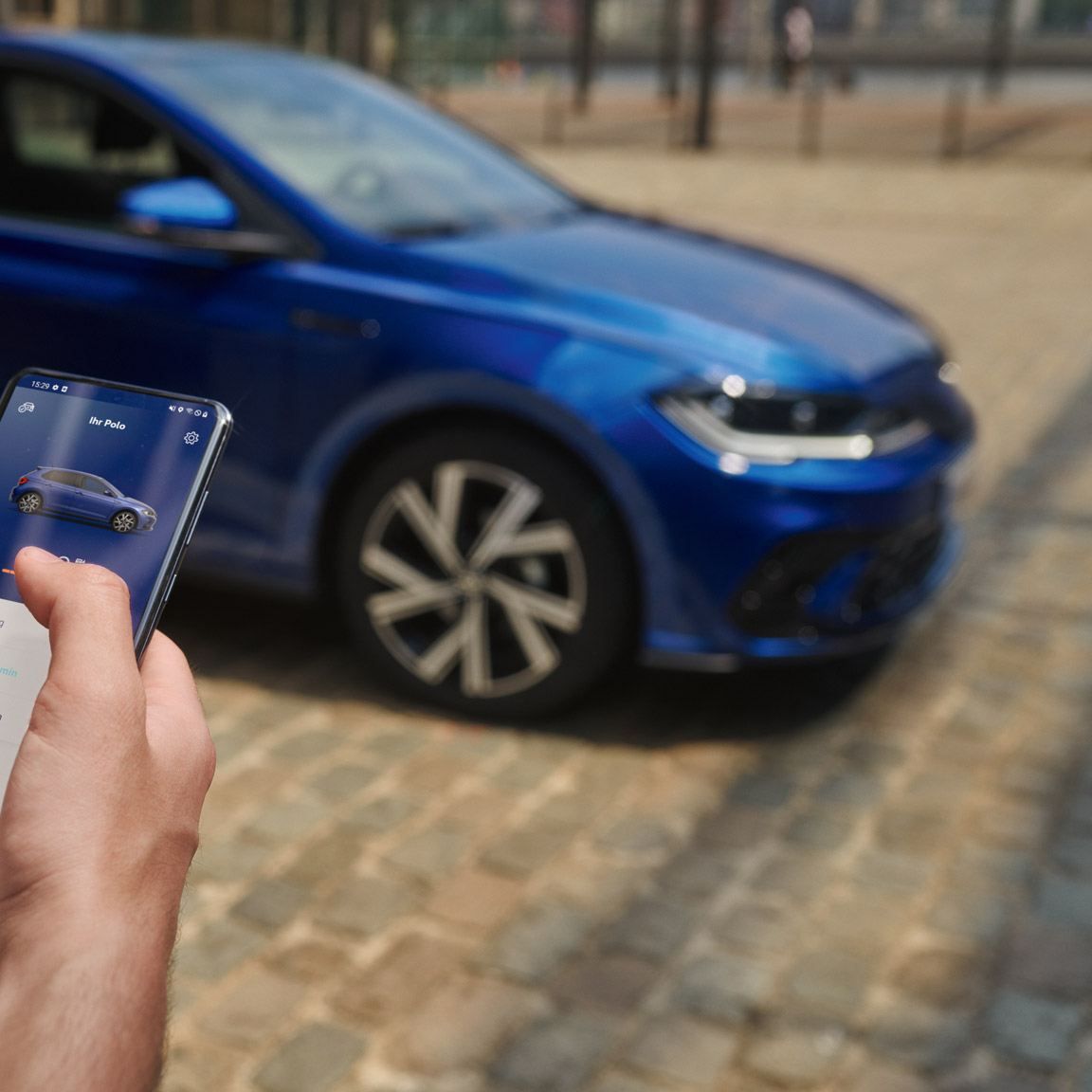 Blick auf ein Handydisplay mit Fahrzeugdaten des VW Polo, im Hintergrund der VW Polo in blau, parkend.