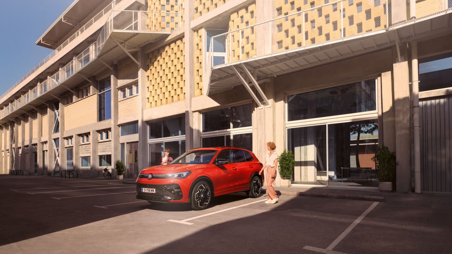 Eine Frau und ein Junge gehen auf einen roten VW Tiguan R-Line zu, der vor einem modernen Gebäude steht.