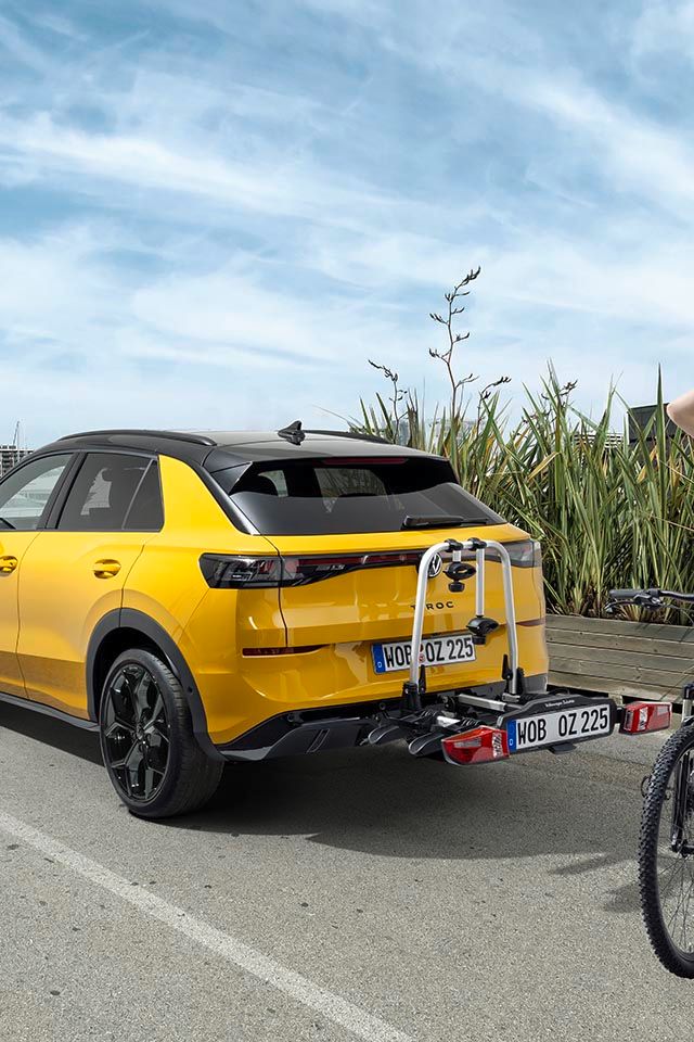 Eine Person mit einem Fahrrad steht neben einem gelben Volkswagen T-Roc mit Fahrradheckträger.