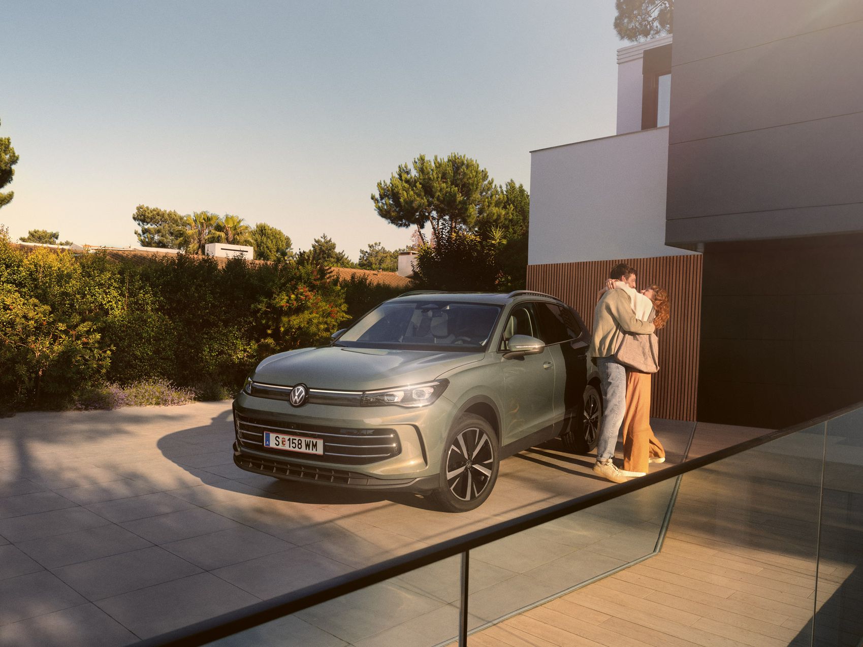 Eine Frau und ein Mann umarmen sich, neben ihnen steht ein VW Tiguan Elegance in der Farbe Cipressino-Grün Metallic