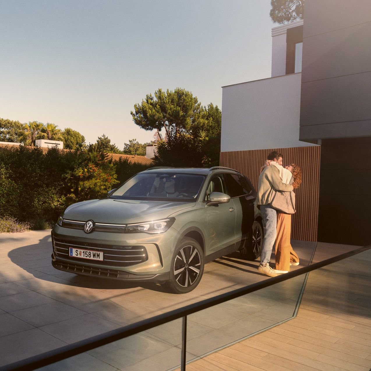 Eine Frau und ein Mann umarmen sich, neben ihnen steht ein VW Tiguan Elegance in der Farbe Cipressino-Grün Metallic