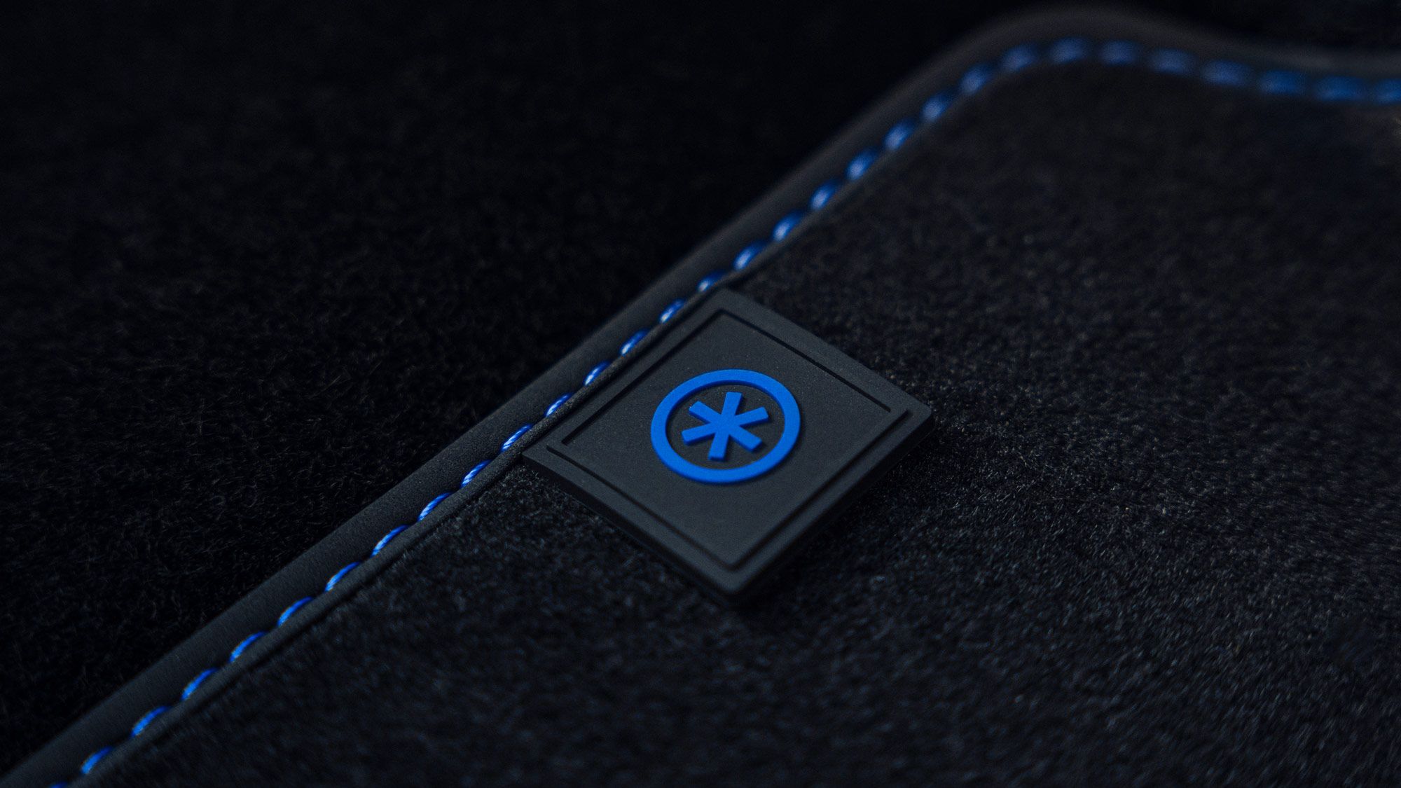 Nahaufnahme einer schwarzen Textiloberfläche mit blauen Nähten. In der Mitte ist ein quadratisches Emblem mit einem blauen Schneesymbol auf schwarzem Hintergrund zu sehen.