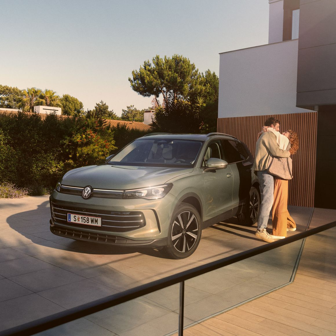 Eine Frau und ein Mann umarmen sich, neben ihnen steht ein VW Tiguan Elegance in der Farbe Cipressino-Grün Metallic