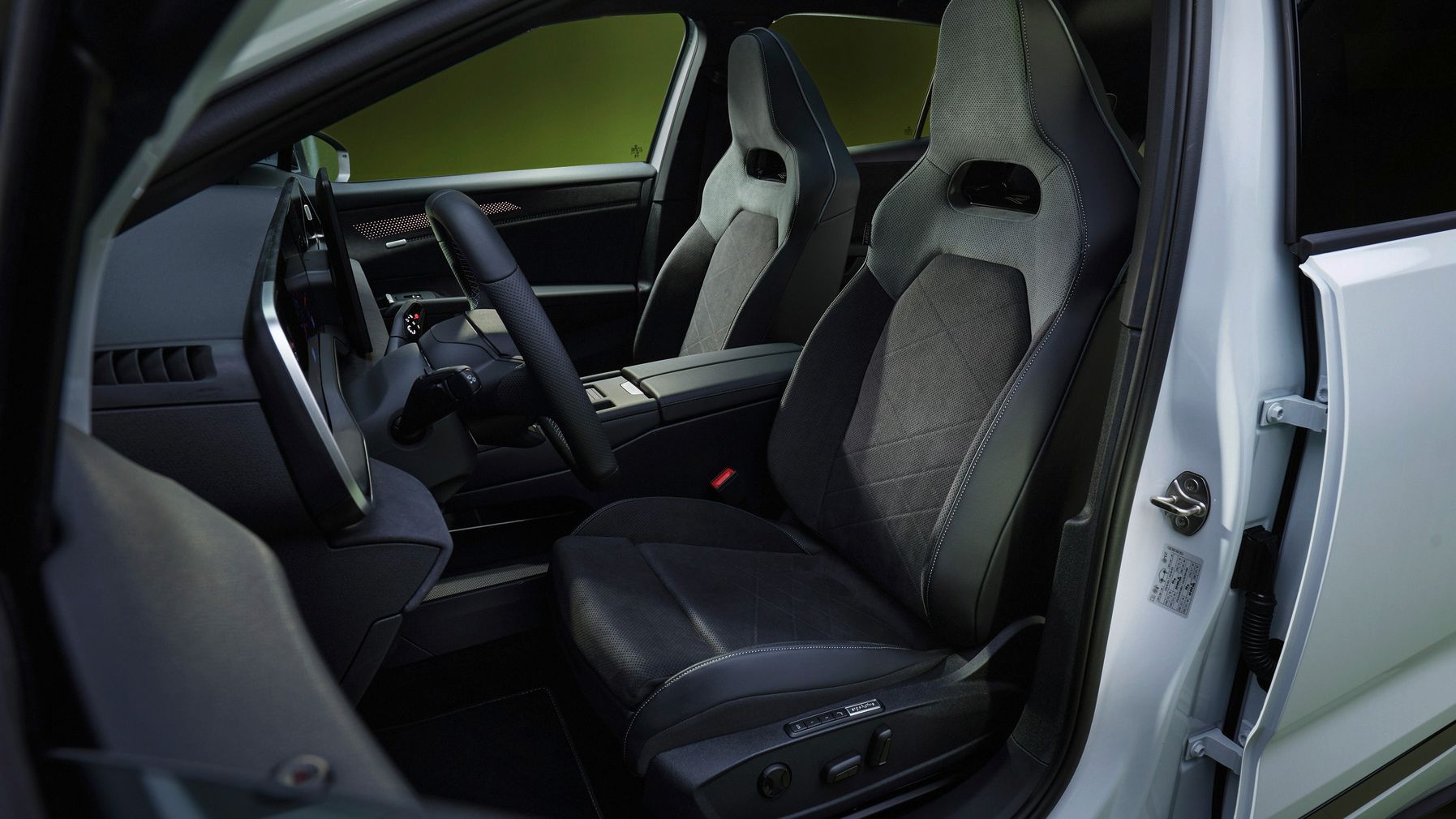 Innenraum eines modernen Autos mit Fokus auf den beiden Vordersitzen in schwarzem Alcantara oder Leder. Man sieht ein sportliches, minimalistisches Cockpit mit breiter Mittelkonsole und wenigen sichtbaren Bedienelementen. Die Tür ist geöffnet, sodass Sitzverstellung und Materialdetails gut erkennbar sind.
