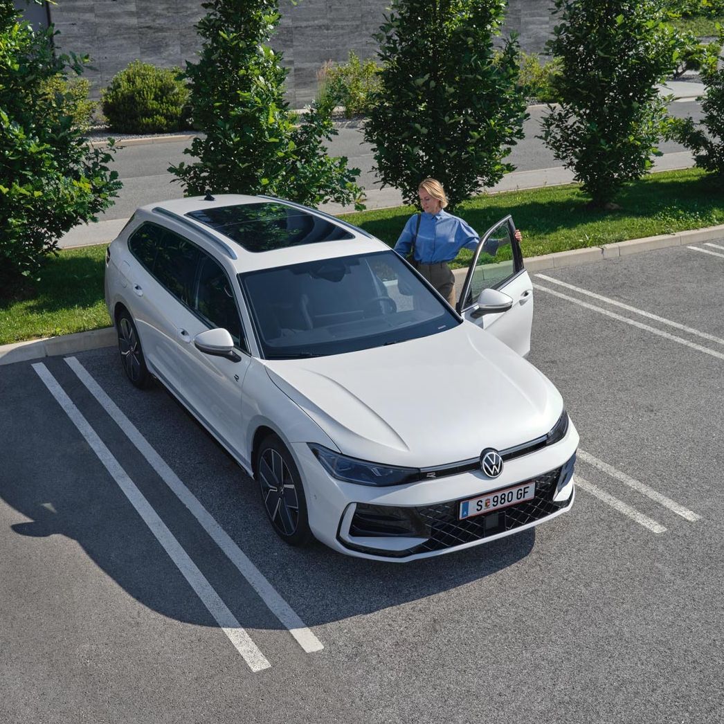 Topshot eines weißen VW Passat R-Line auf einem Parkplatz, eine Frau steigt auf der Fahrerseite ein.