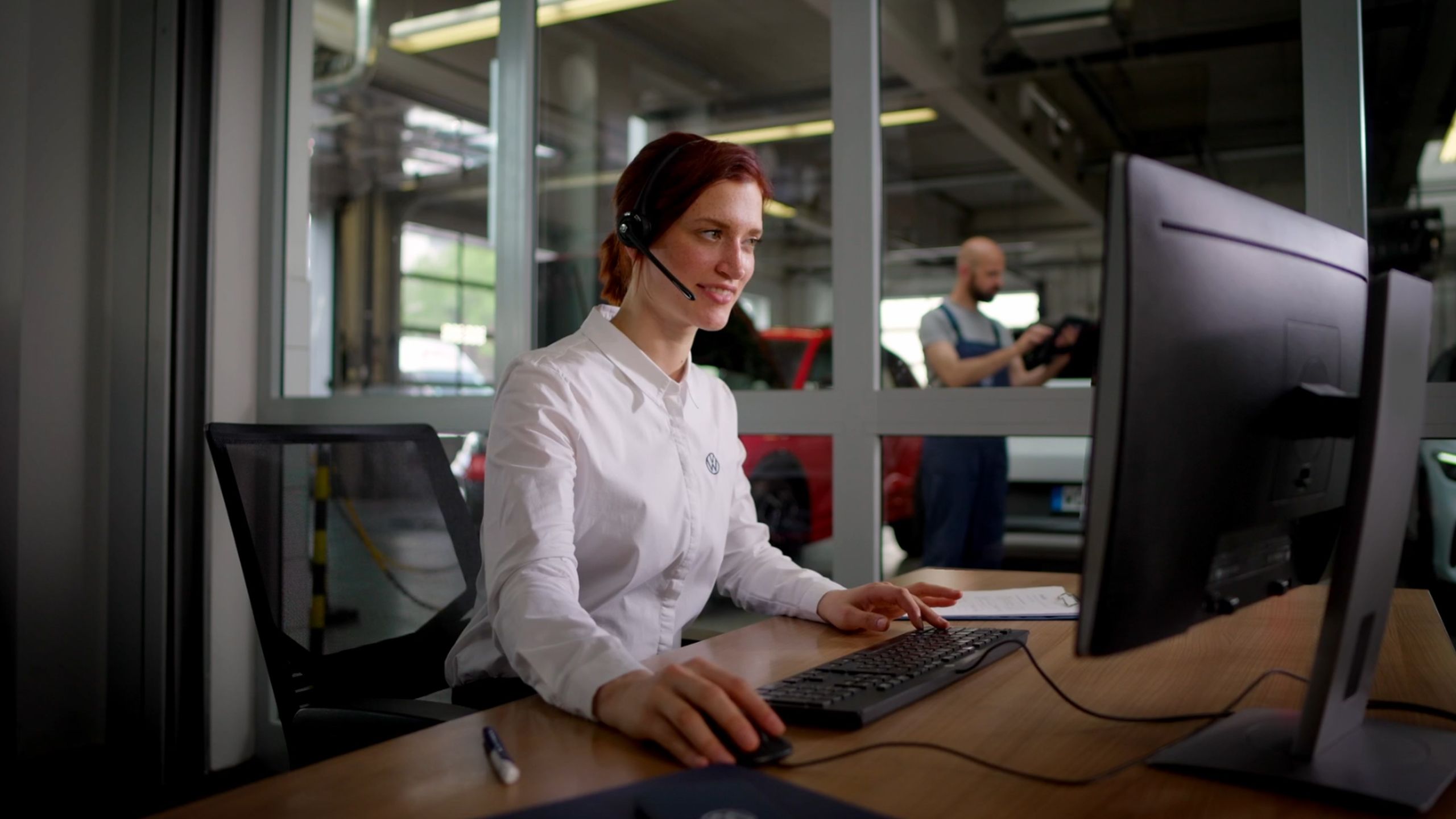Eine Frau mit Kopfhörern arbeitet an einem Computer. Im Hintergrund ist eine Werkstatt mit einem Mechaniker zu sehen.