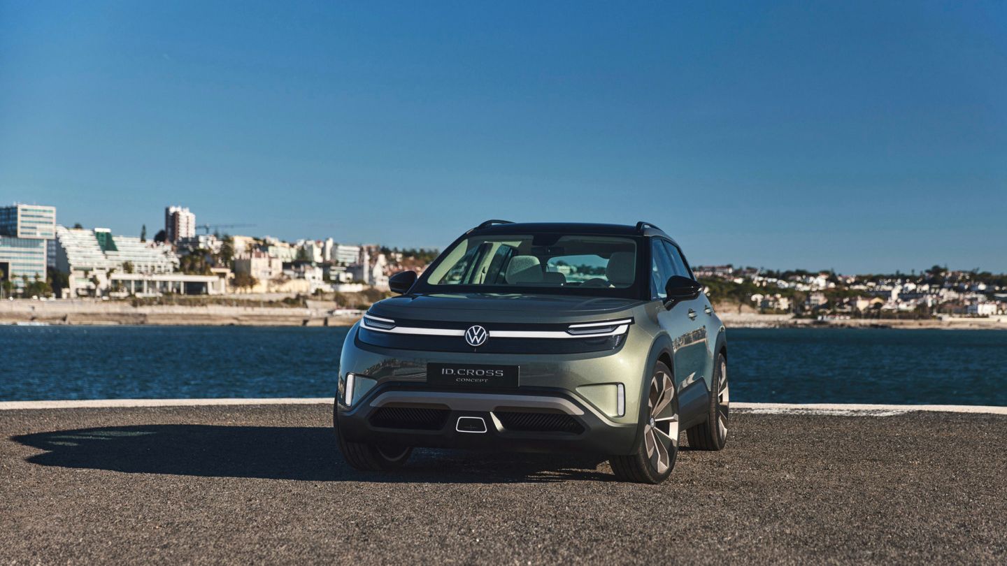 Ein grüner Volkswagen ID. Cross steht vor einem Wasserhintergrund mit Stadtansicht. Das Auto ist von vorne gesehen und strahlt in der Sonne. Es gibt keine anderen Fahrzeuge oder Personen im Bild.