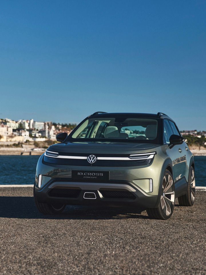 Ein grüner Volkswagen ID. Cross steht vor einem Wasserhintergrund mit Stadtansicht. Das Auto ist von vorne gesehen und strahlt in der Sonne. Es gibt keine anderen Fahrzeuge oder Personen im Bild.