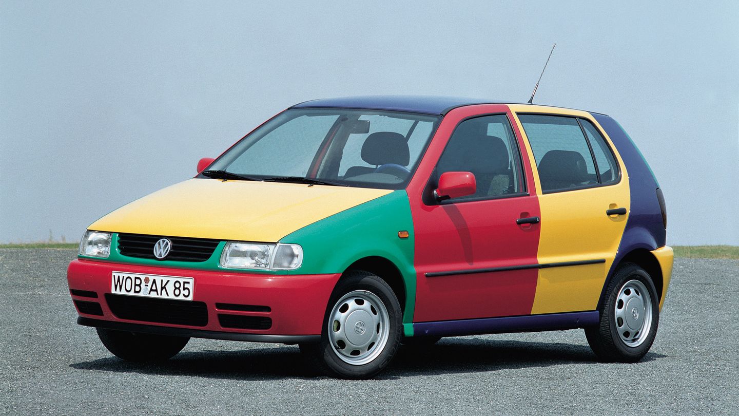 Ein VW Polo Harlekin mit einer bunten Lackierung in Rot, Gelb, Grün und Blau steht auf einer Straße. Das Auto hat eine vierseitige Farbteilung, die jedem Teil eine unterschiedliche Farbe gibt. Im Hintergrund ist ein bewölkter Himmel und grünes Gras zu sehen.
