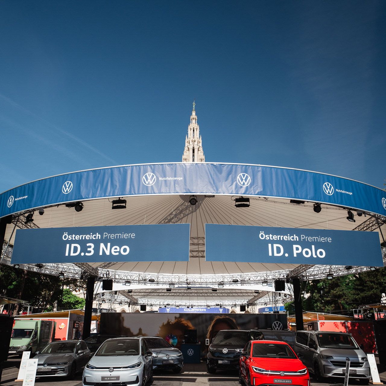 Outdoor-Ausstellung von Volkswagen mit mehreren geparkten Autos vor einer runden, überdachten Bühne. Auf großen blauen Bannern stehen die Texte „Österreich Premiere ID.3 Neo“ und „ID. Polo“. Im Hintergrund ragt ein historischer Turm in den blauen Himmel.
