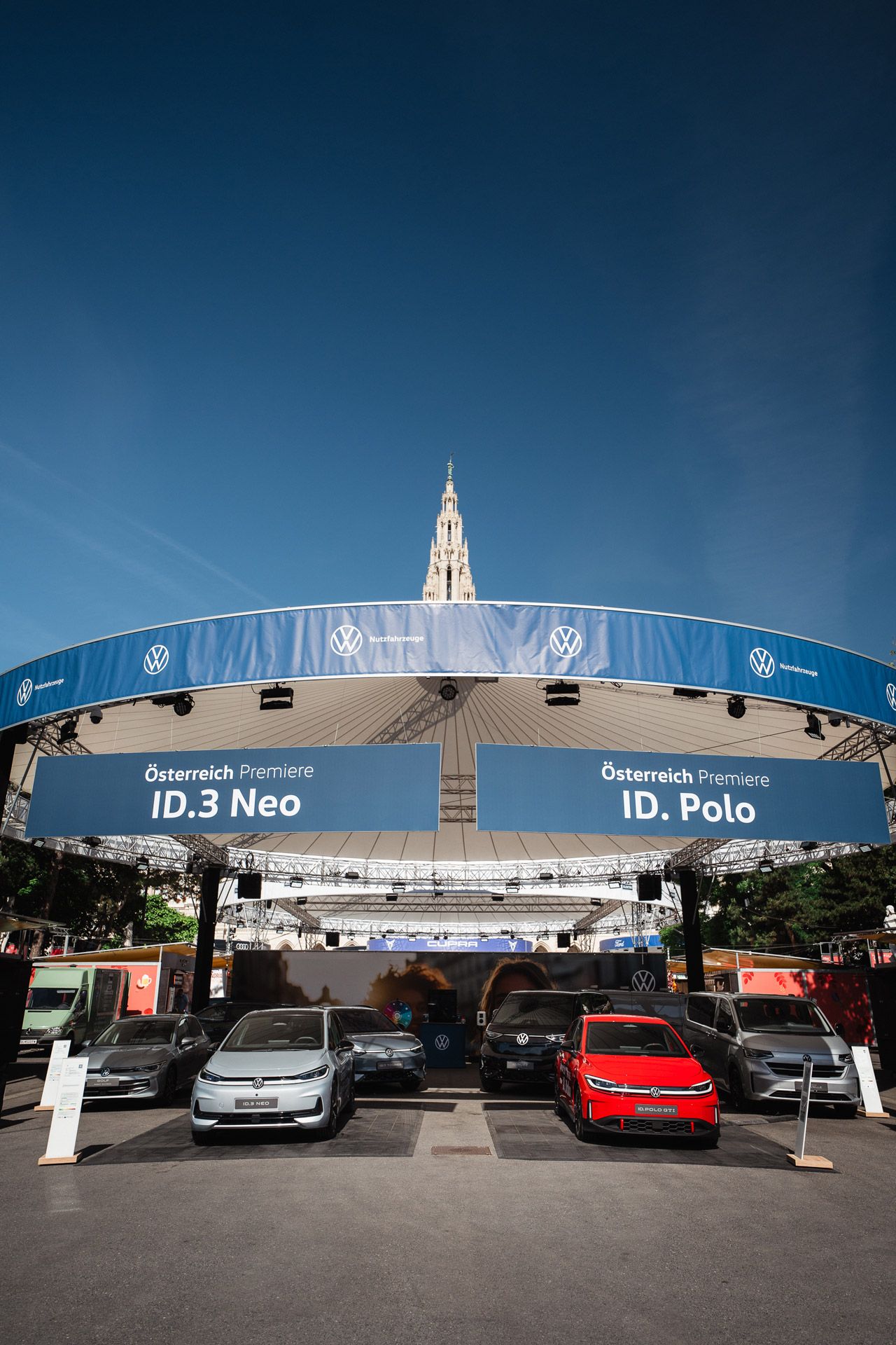 Outdoor-Ausstellung von Volkswagen mit mehreren geparkten Autos vor einer runden, überdachten Bühne. Auf großen blauen Bannern stehen die Texte „Österreich Premiere ID.3 Neo“ und „ID. Polo“. Im Hintergrund ragt ein historischer Turm in den blauen Himmel.