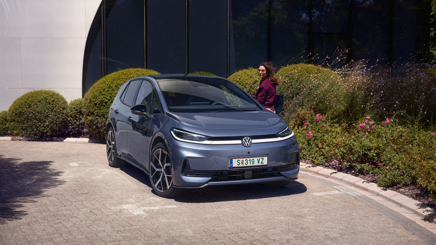 Eine Frau steht an der Fahrertür eines blauen VW ID.3 Neo, der vor einem modernen Bürogebäude parkt