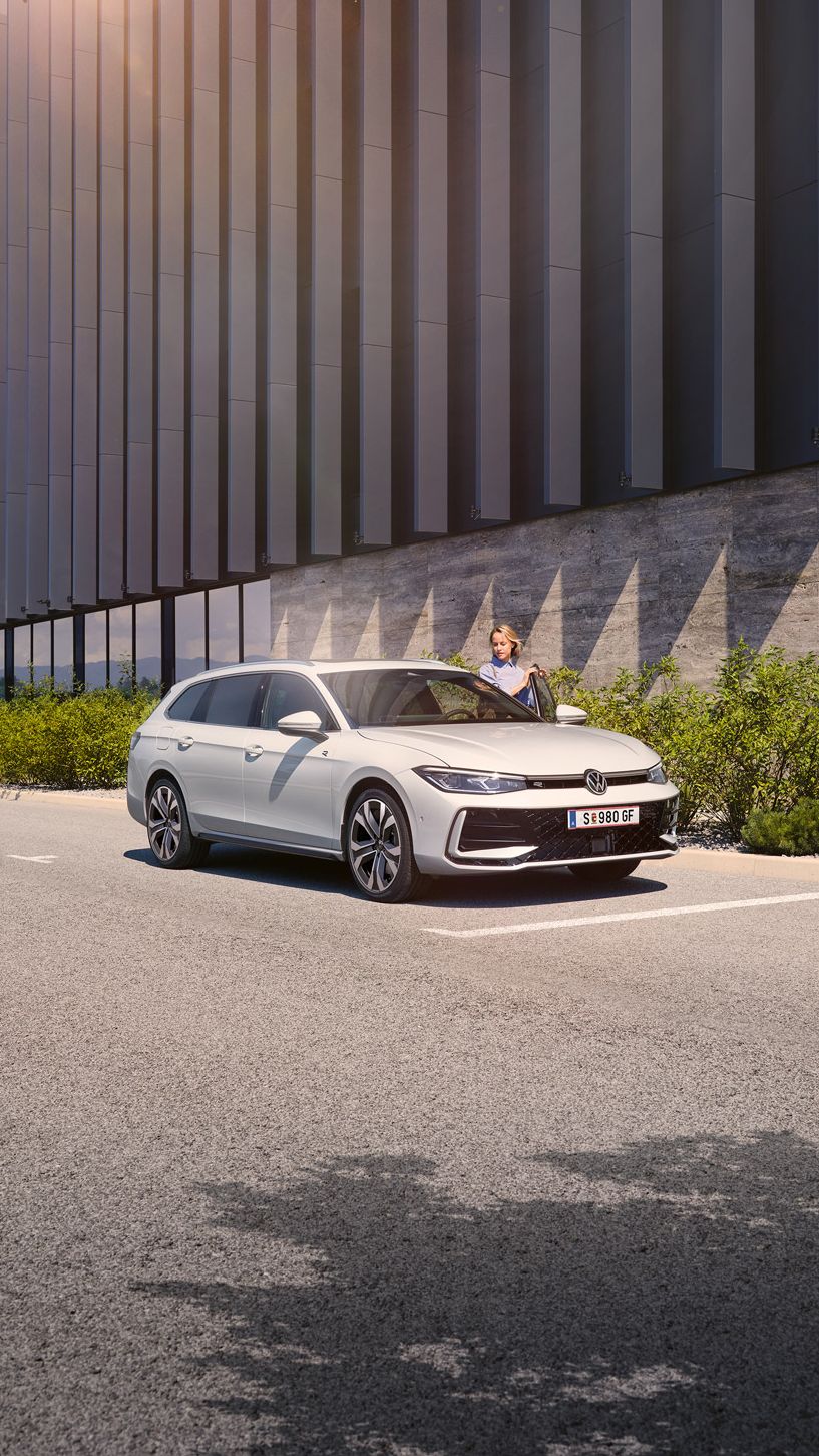  Eine Frau öffnet die Fahrertür eines weißen VW Passat R-Line und steigt ein. Im Hintergrund sieht man ein Gebäude.