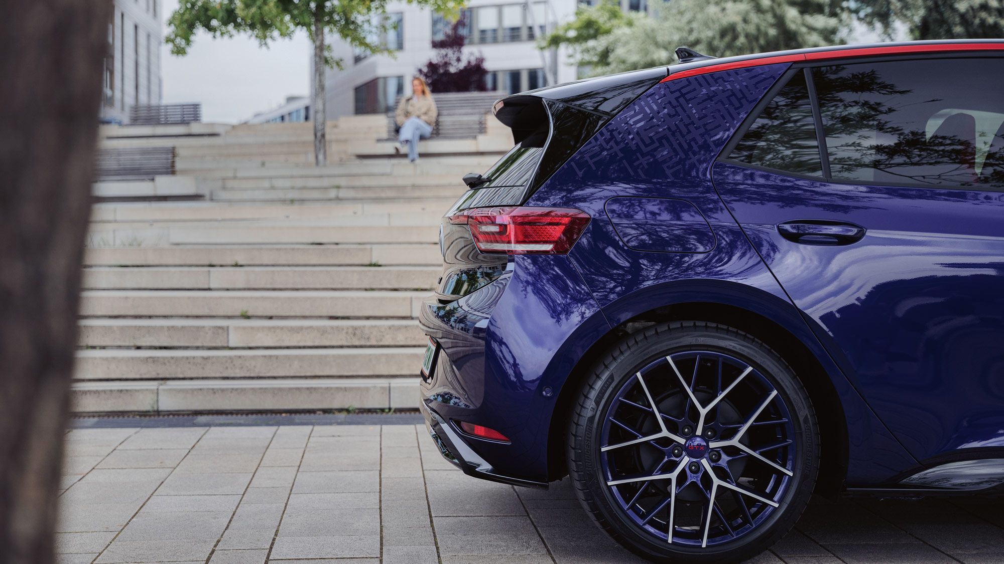 Ein blauer Kleinwagen ist auf einem gepflasterten Platz geparkt. Im Hintergrund ist eine Treppe mit einer Person, die darauf sitzt. Ein Baum ragt links ins Bild.