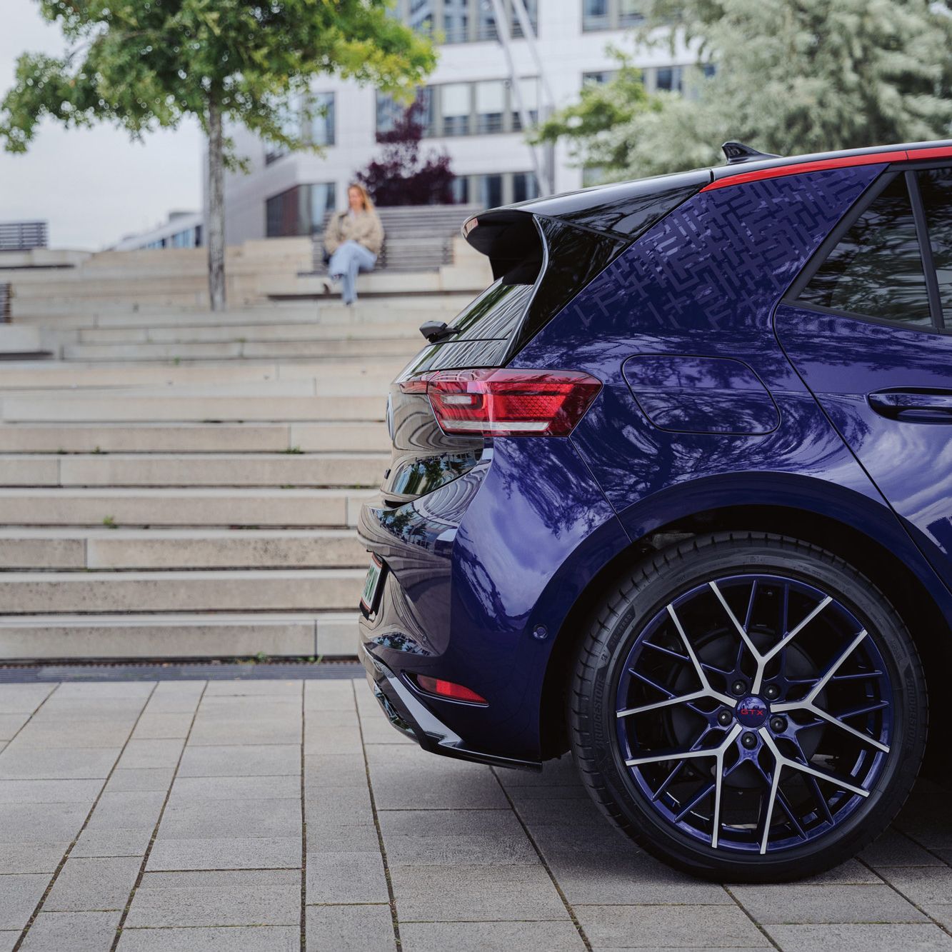 Ein blauer ID.3 GTX FIRE+ICE ist auf einem gepflasterten Platz geparkt. Im Hintergrund ist eine Treppe mit einer Person, die darauf sitzt. Ein Baum ragt links ins Bild.