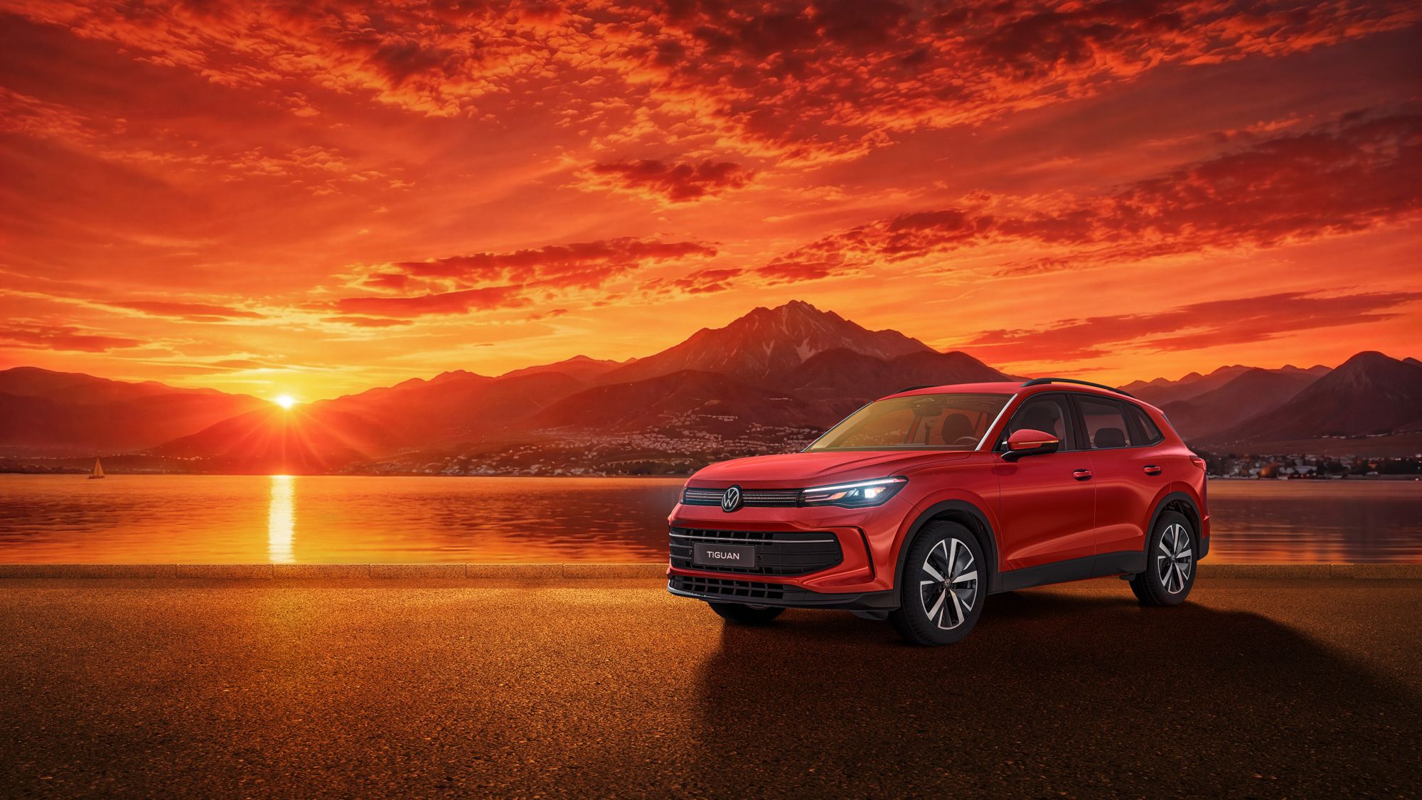 Roter Tiguan auf einer glatten Uferstraße vor einem See bei Sonnenuntergang. Im Hintergrund erheben sich dunkle Bergsilhouetten unter einem dramatisch orange-roten Himmel mit dichten Wolken. Das Auto steht rechts im Bild und wirkt modern und glänzend, während die Sonne links knapp über dem Horizont steht.