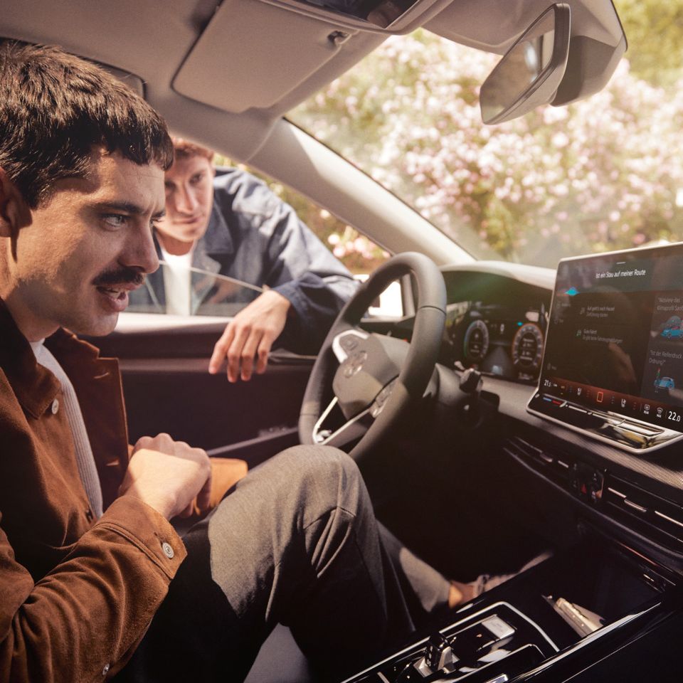 Ein Mann sitzt im neuen VW Golf und schaut auf das Infotainment-Display, ein anderer schaut durch das Fenster.
