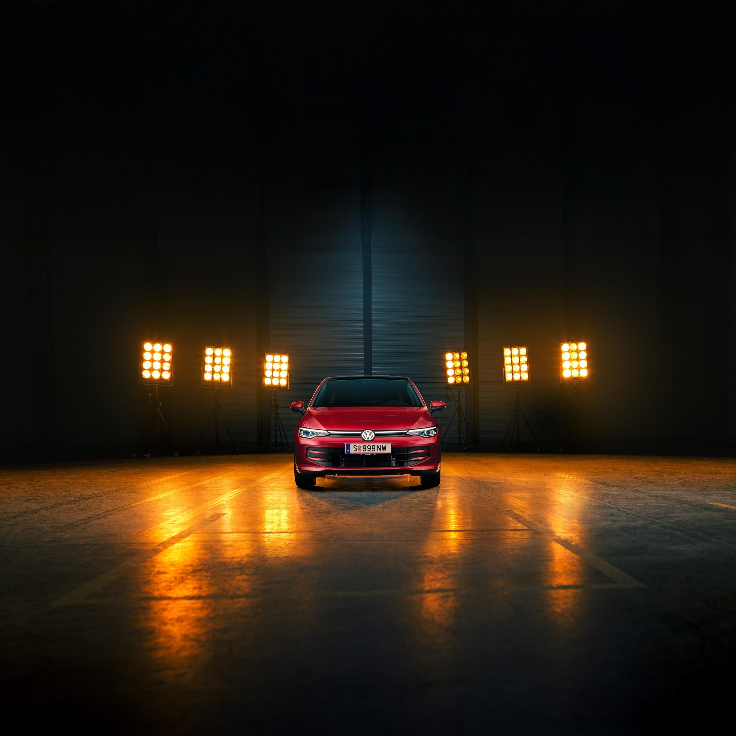 Ein roter VW Golf Falco Edition in Frontalansicht steht mittig im Bild in einer leeren Halle, Bühnenlichter strahlen das Fahrzeug von hinten an.