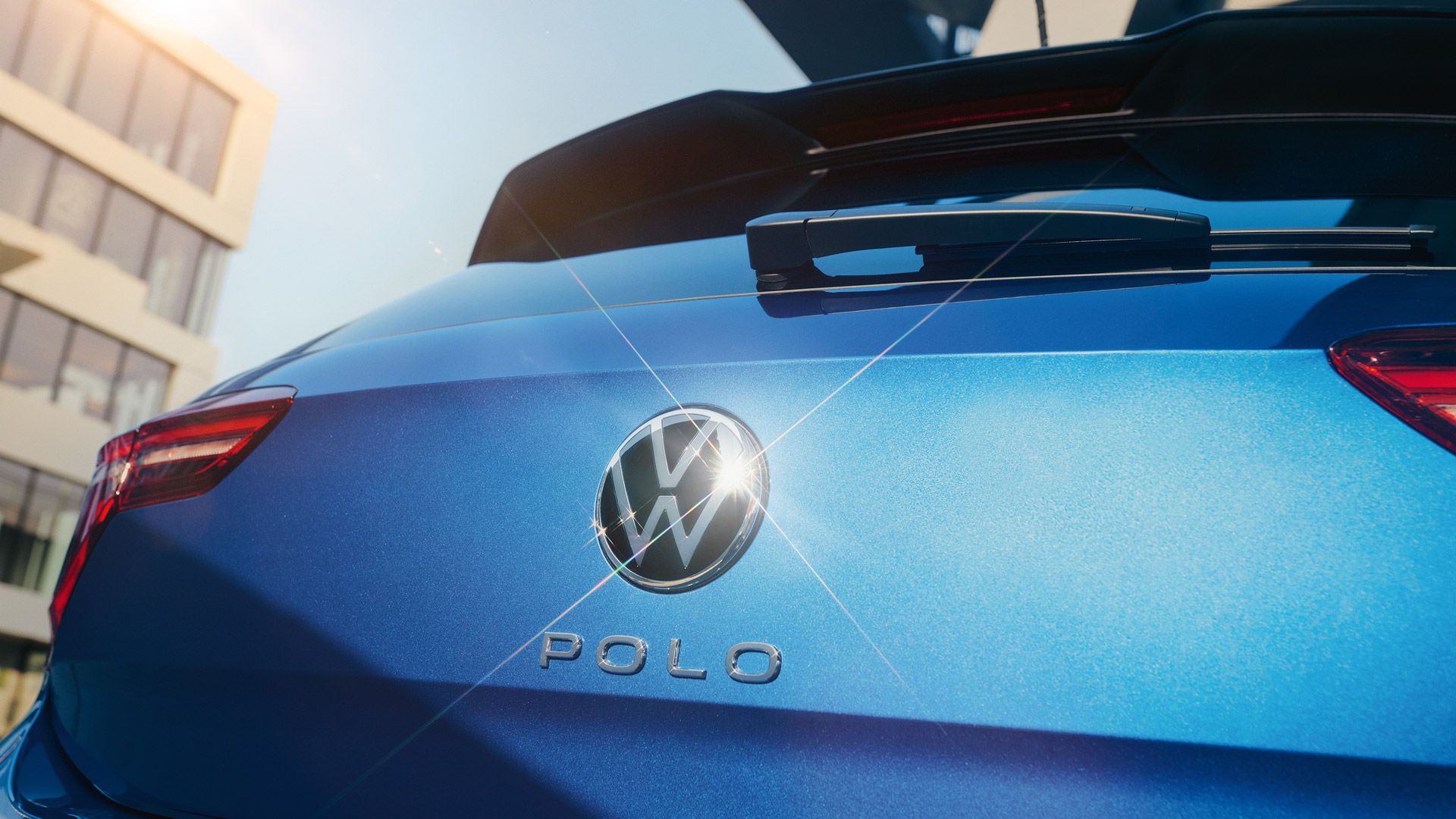 Die geschlossene Kofferraumklappe eines blauen VW Polo mit VW-Logo, in dem sich die Sonne spiegelt.