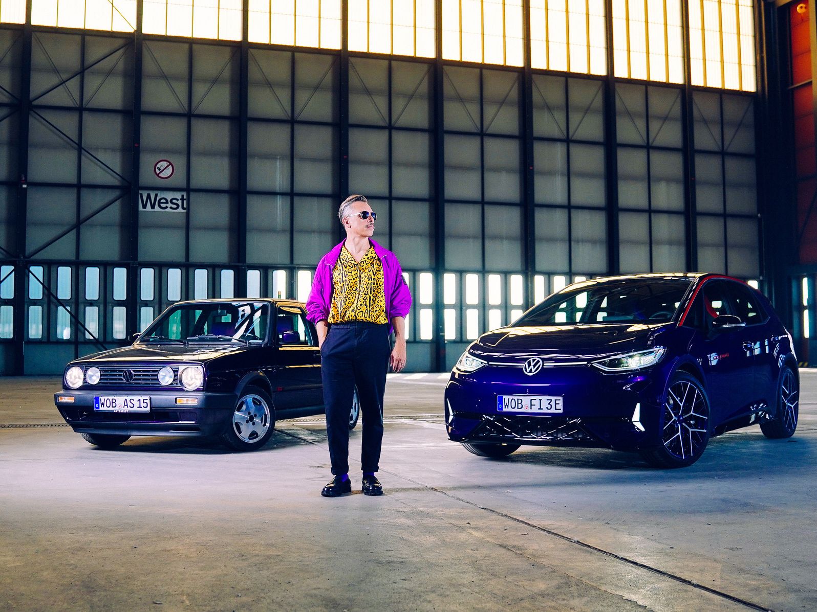 Eine Person steht in einer großen Halle zwischen einem schwarzen und einem blauen Auto. Die Autos sind von der Marke Volkswagen. Die Halle ist größtenteils leer und die Wände bestehen aus vielen Fensterpaneelen.