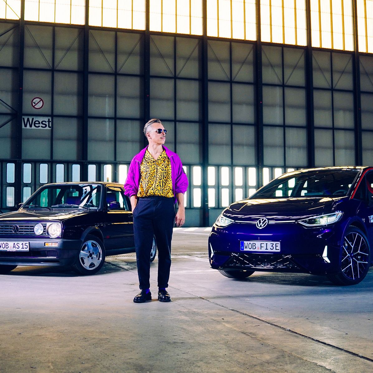 Eine Person steht in einer großen Halle zwischen einem schwarzen und einem blauen Auto. Die Autos sind von der Marke Volkswagen. Die Halle ist größtenteils leer und die Wände bestehen aus vielen Fensterpaneelen.