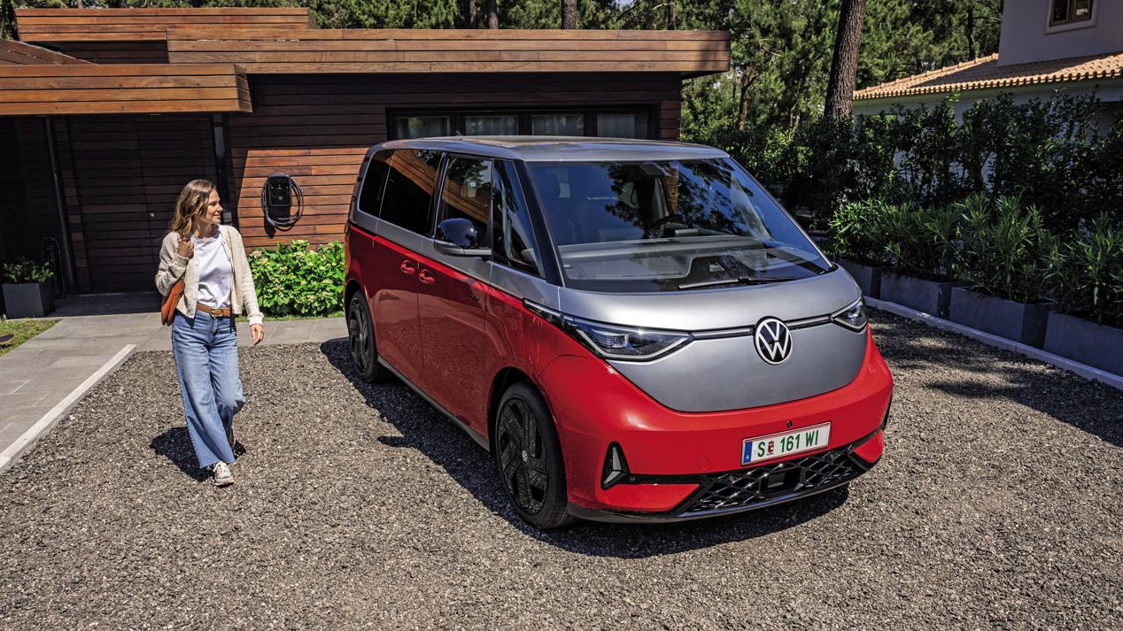 Eine Person steht neben einem roten Volkswagen ID. Buzz GTX, der in einer gepflasterten Einfahrt vor einem modernen Haus geparkt ist.