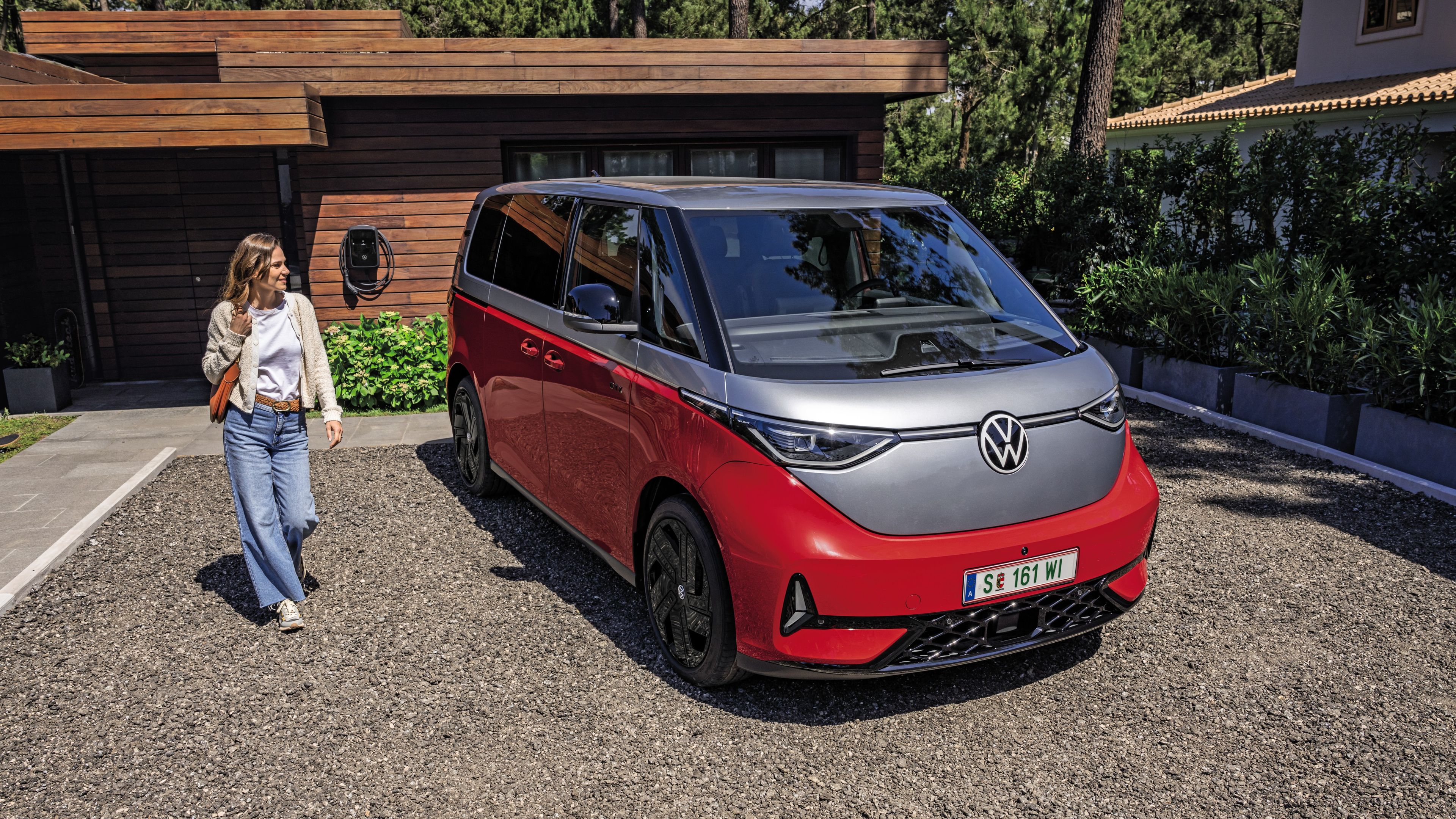 Eine Person steht neben einem roten Volkswagen ID. Buzz GTX, der in einer gepflasterten Einfahrt vor einem modernen Haus geparkt ist.