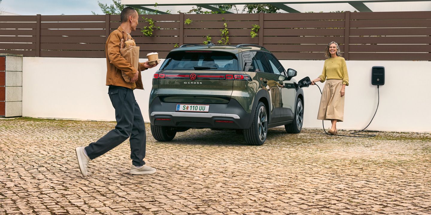 Eine Frau steckt ein Ladekabel einer Wallbox in einen grünen VW ID. Cross, ein Mann geht auf sie zu und hat Kaffeebecher in der Hand und Brot unter dem Arm.